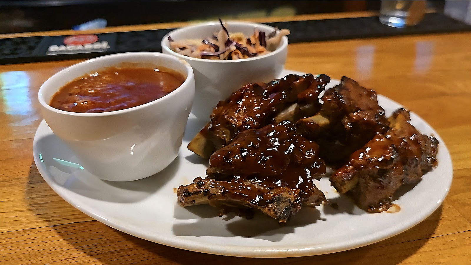 Barbecue-glazed ribs plated with a ramekin of sauce and a bowl of coleslaw.