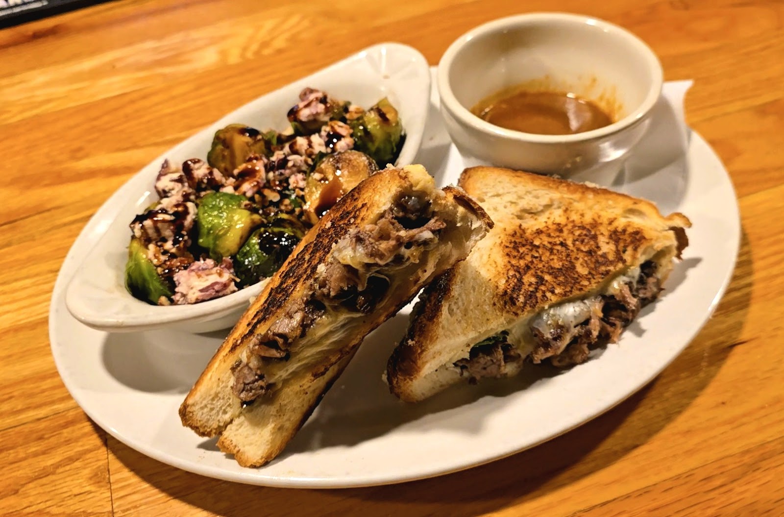 A toasted grilled melt cut in two, next to roasted Brussels sprouts and a dipping cup.
